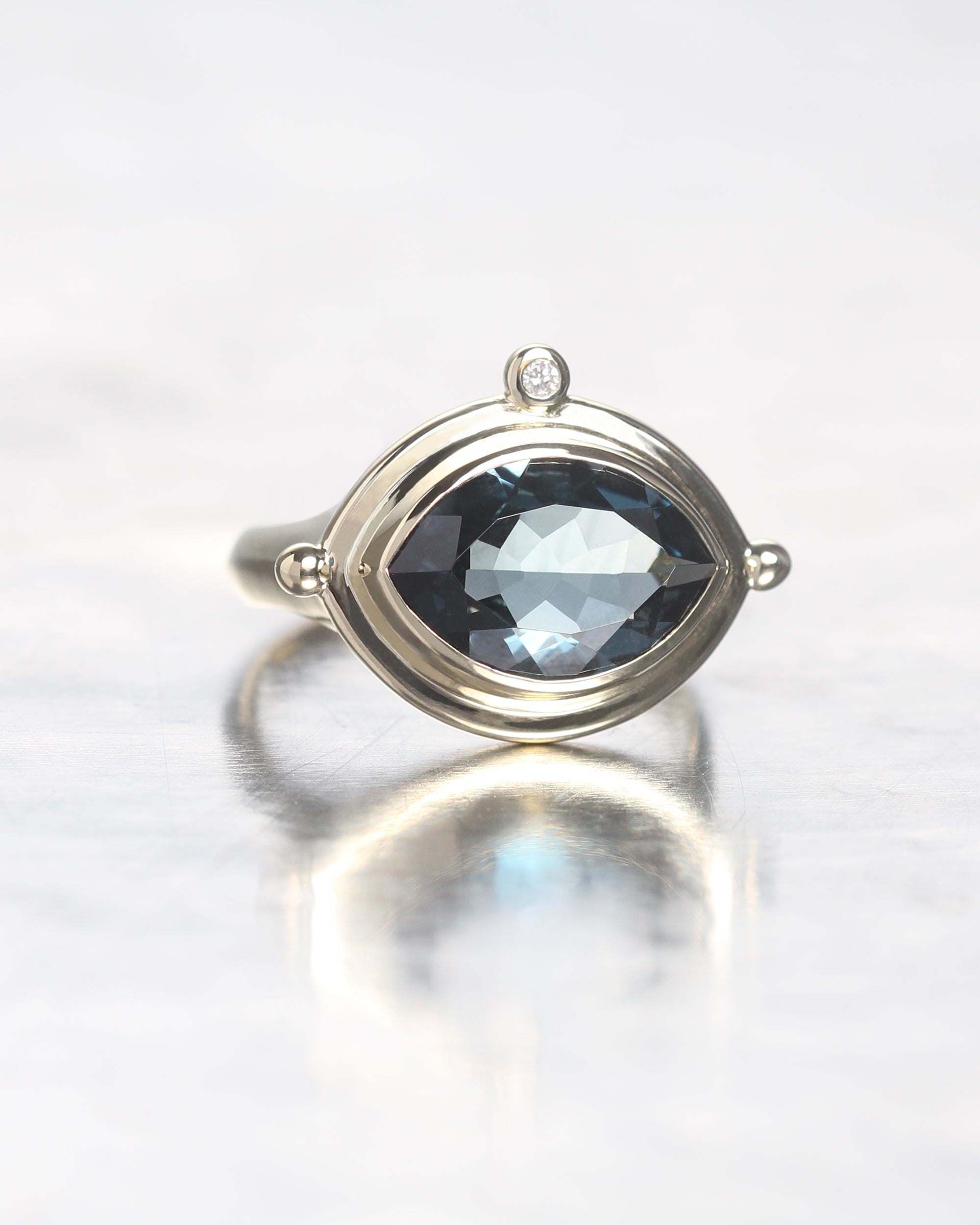 A silver ring featuring a marquise-cut dark blue gemstone in a prong setting, with a small round diamond accent at the top, shown on a reflective light gray surface.