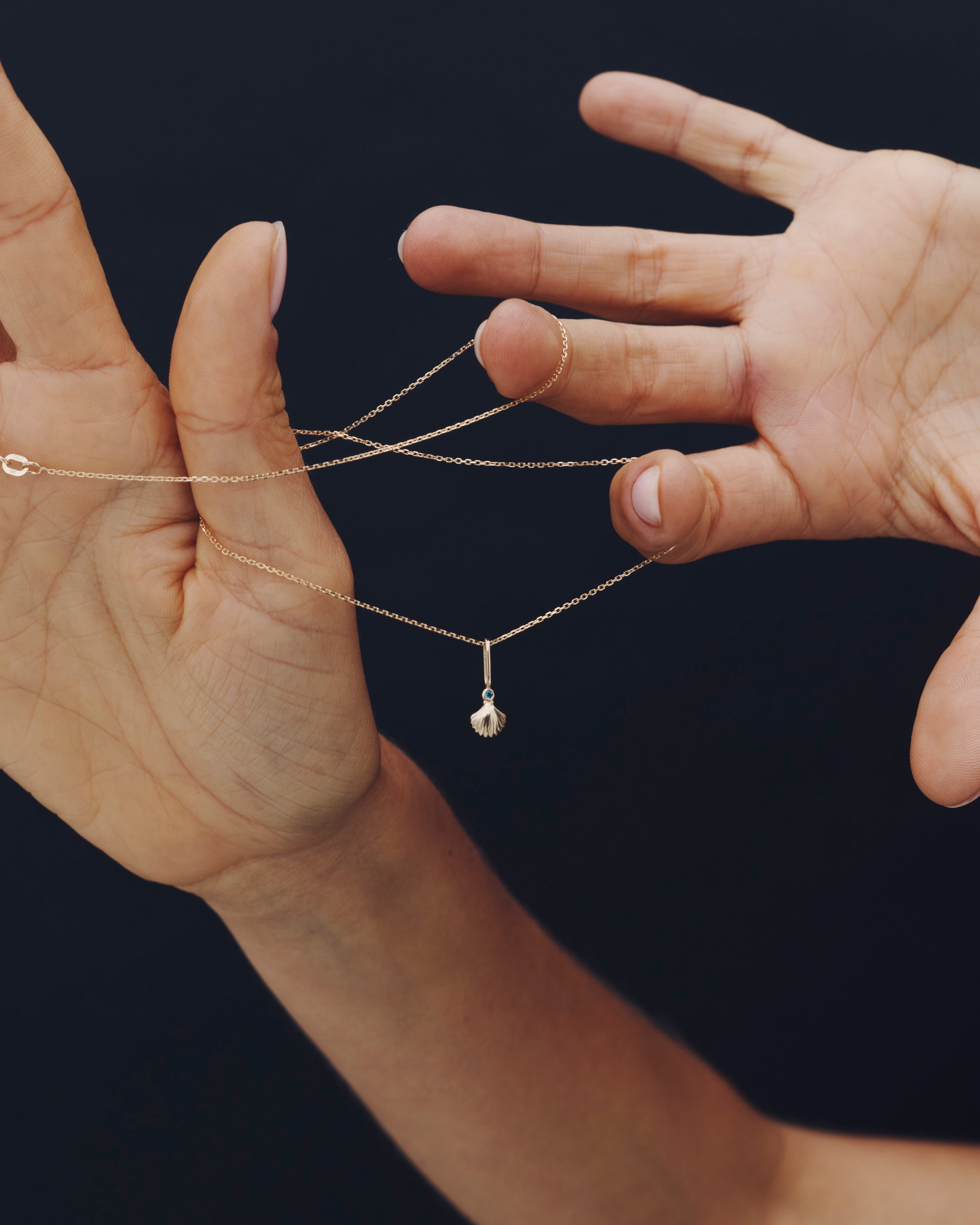 Two hands holding a delicate gold necklace with a pearl pendant against a dark background