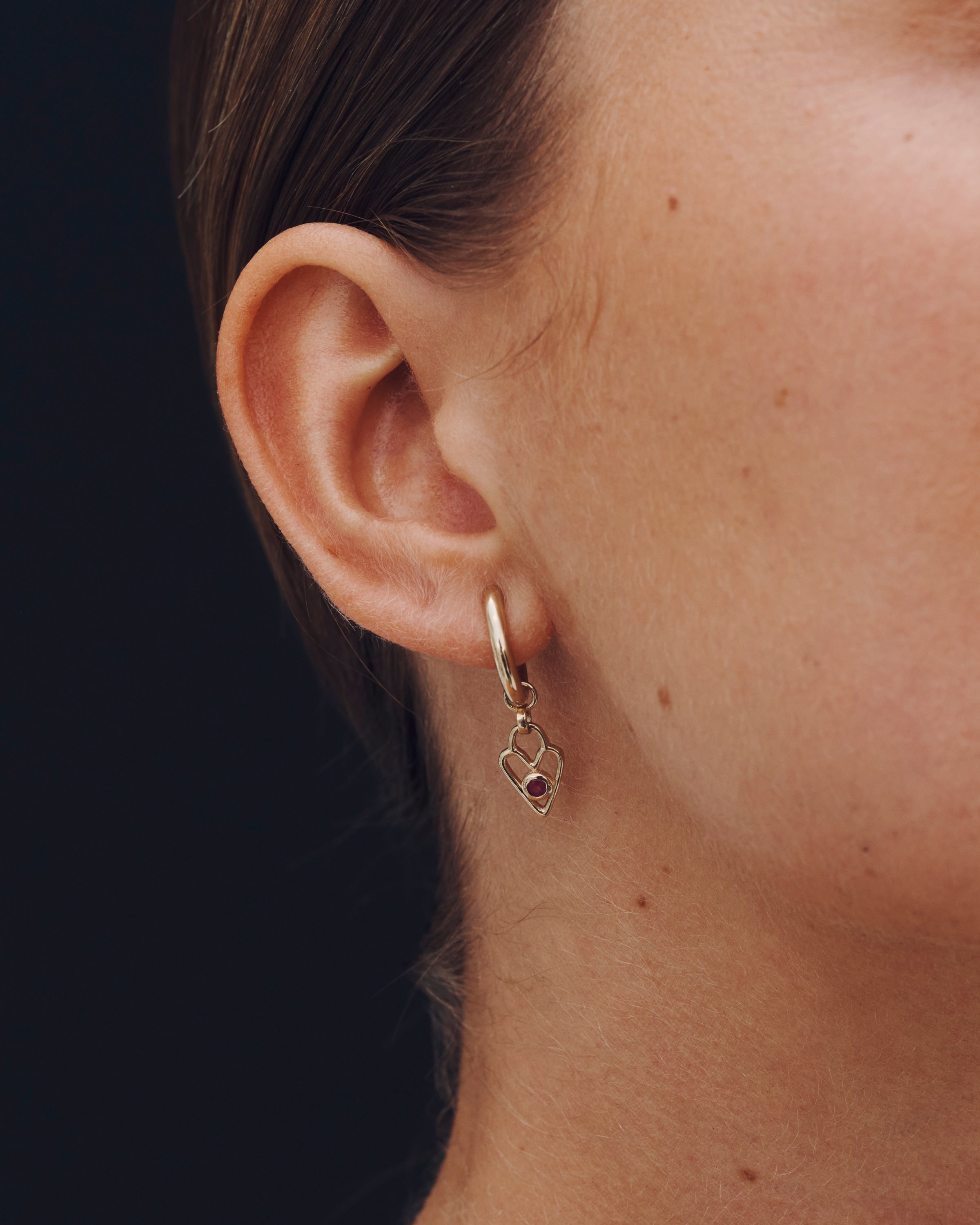 Close-up of an ear wearing a gold hoop earring with a heart charm against a dark background