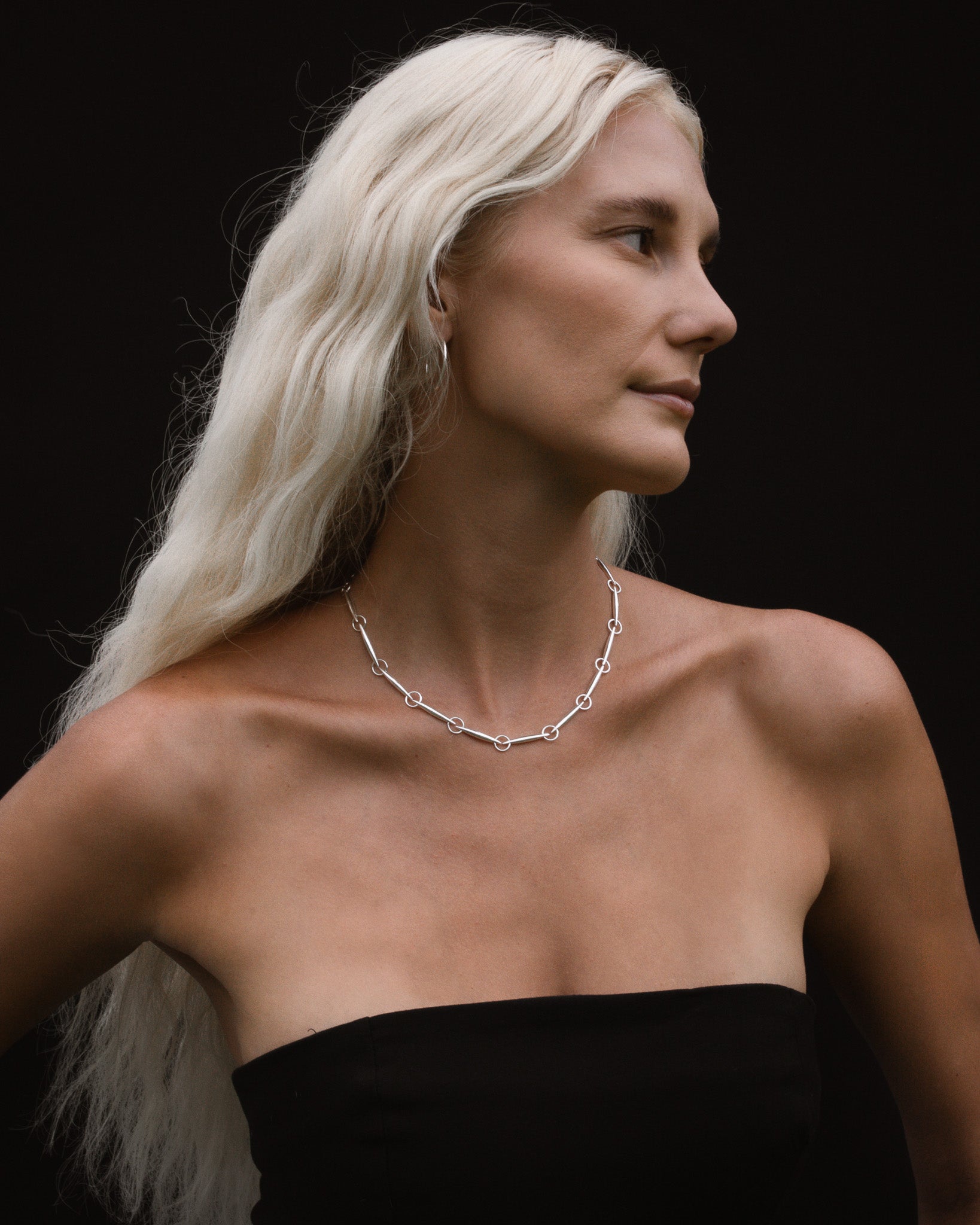 Woman with long white hair wearing a silver necklace against a black background