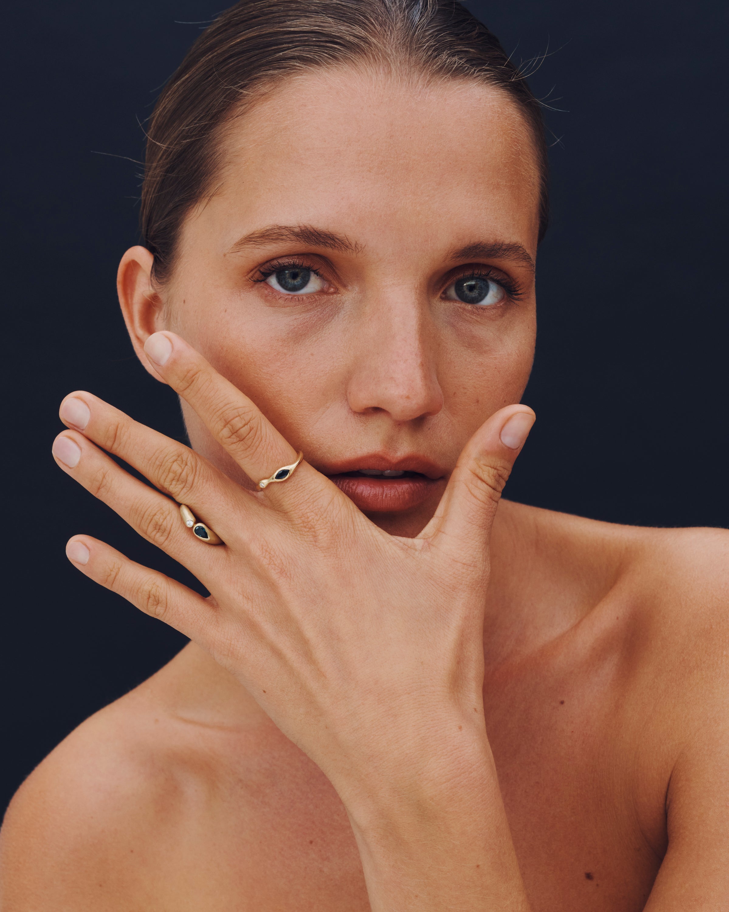 Woman with a hand covering her face, wearing rings on a dark background