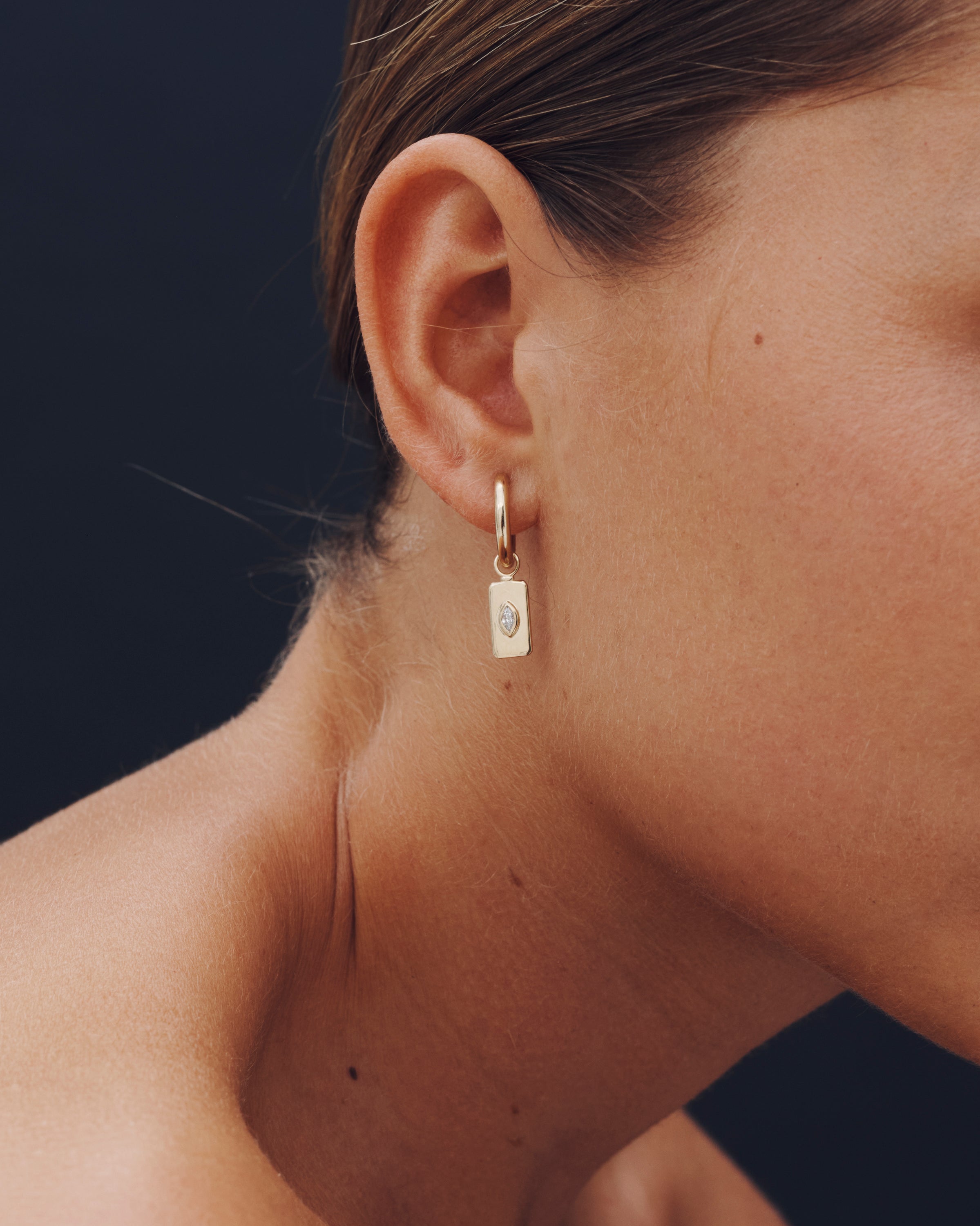 Close-up of a person wearing a gold hoop earring with a small diamond on a dark background