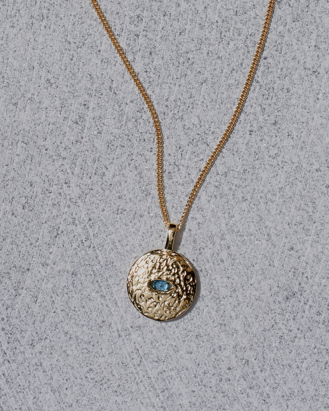 Gold necklace with a textured pendant featuring a blue gemstone on a gray background